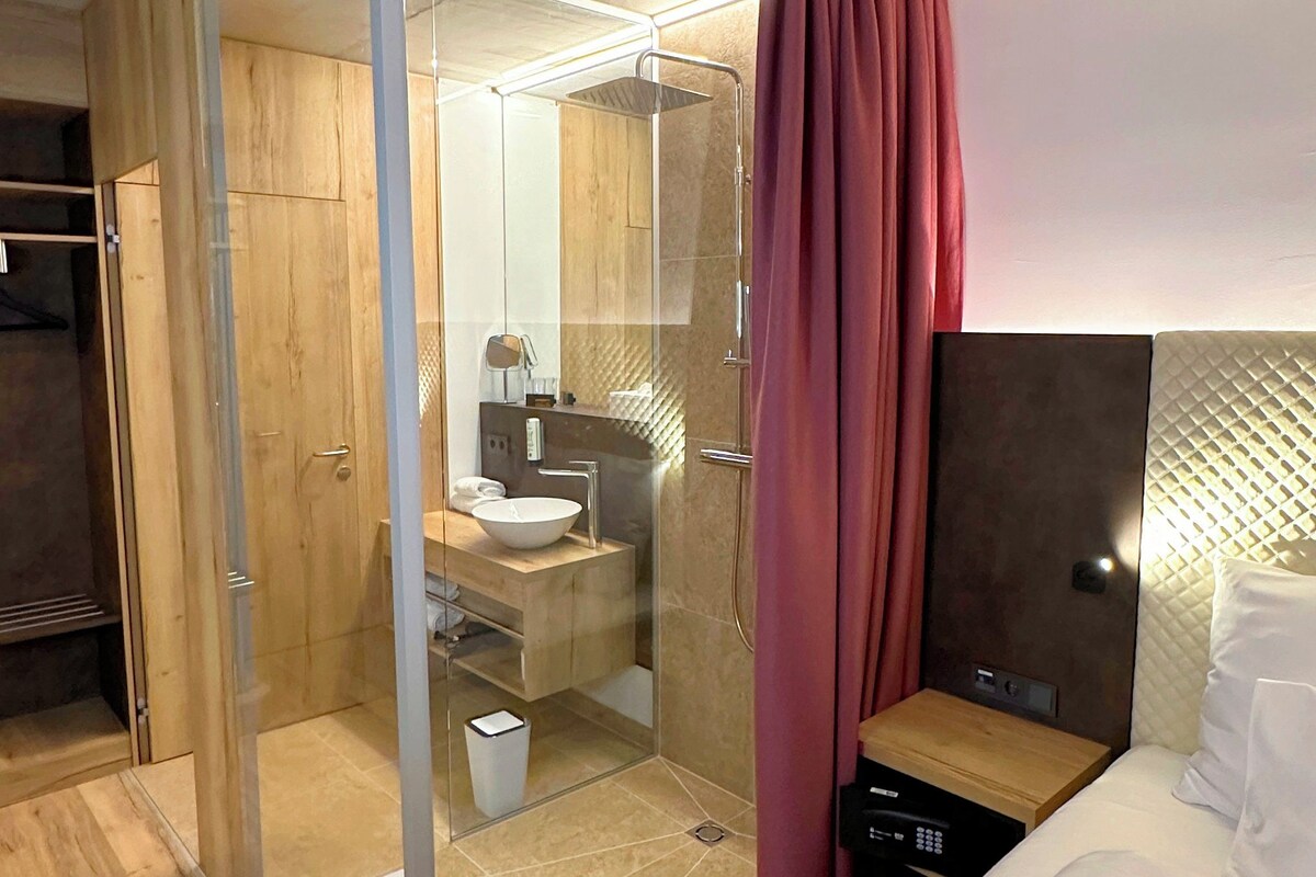 Habitación de hotel moderna con ducha de vidrio, tocador de madera, cama con ropa blanca y cortina roja.