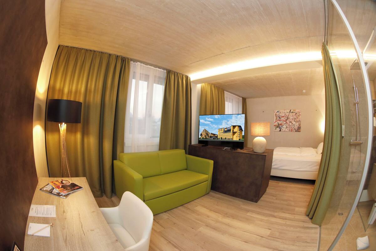 Habitación de hotel con sofá verde, un escritorio de madera con papeles, televisor, cama y cabina de ducha de vidrio.