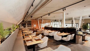 Un restaurant moderne avec de hauts plafonds, des conduits apparents et un espace bar. Les tables sont disposées pour accueillir les convives, et des plantes apportent une touche de verdure.