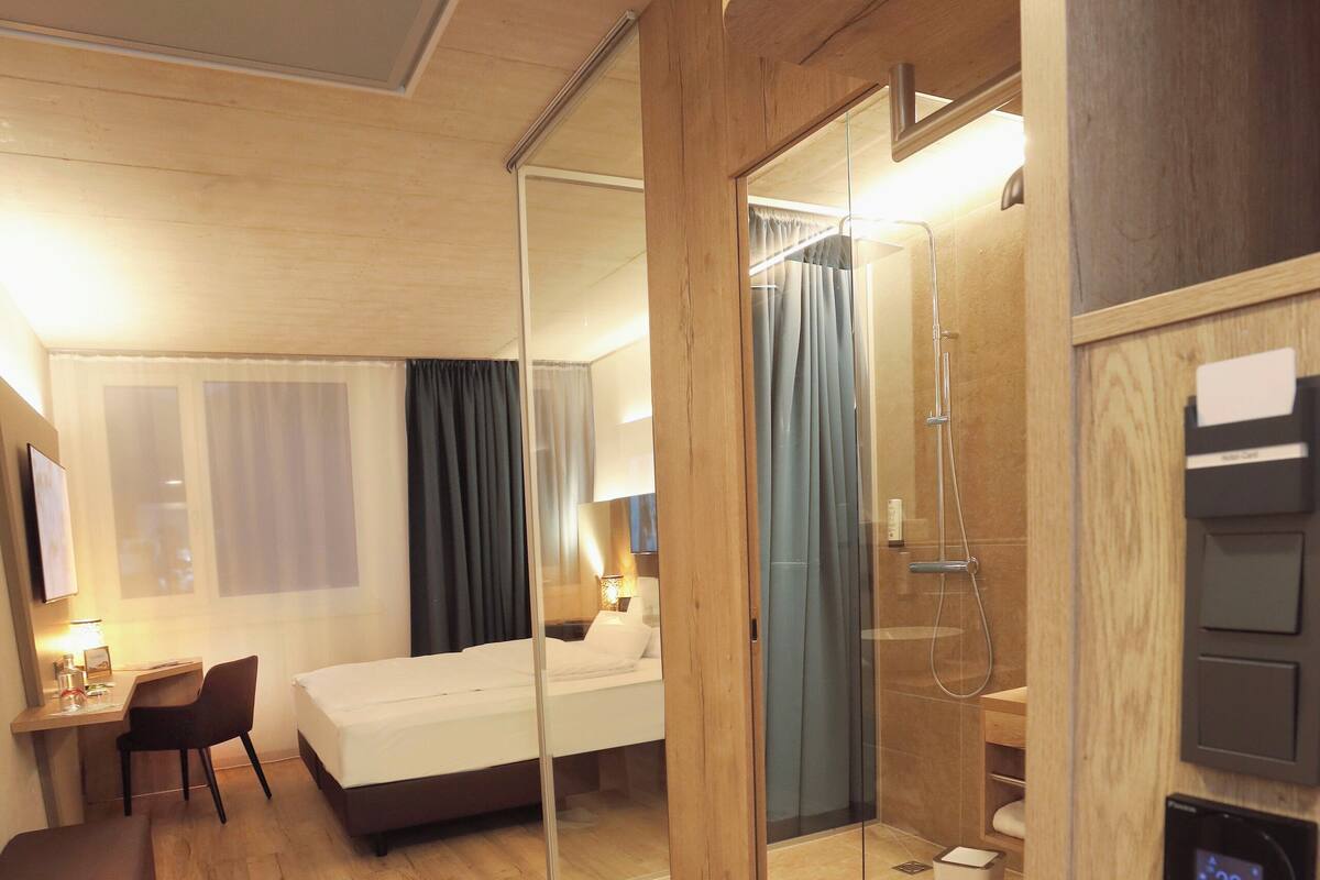 Habitación de hotel con cama, escritorio, silla y un separador de vidrio que delimita la zona de la ducha.