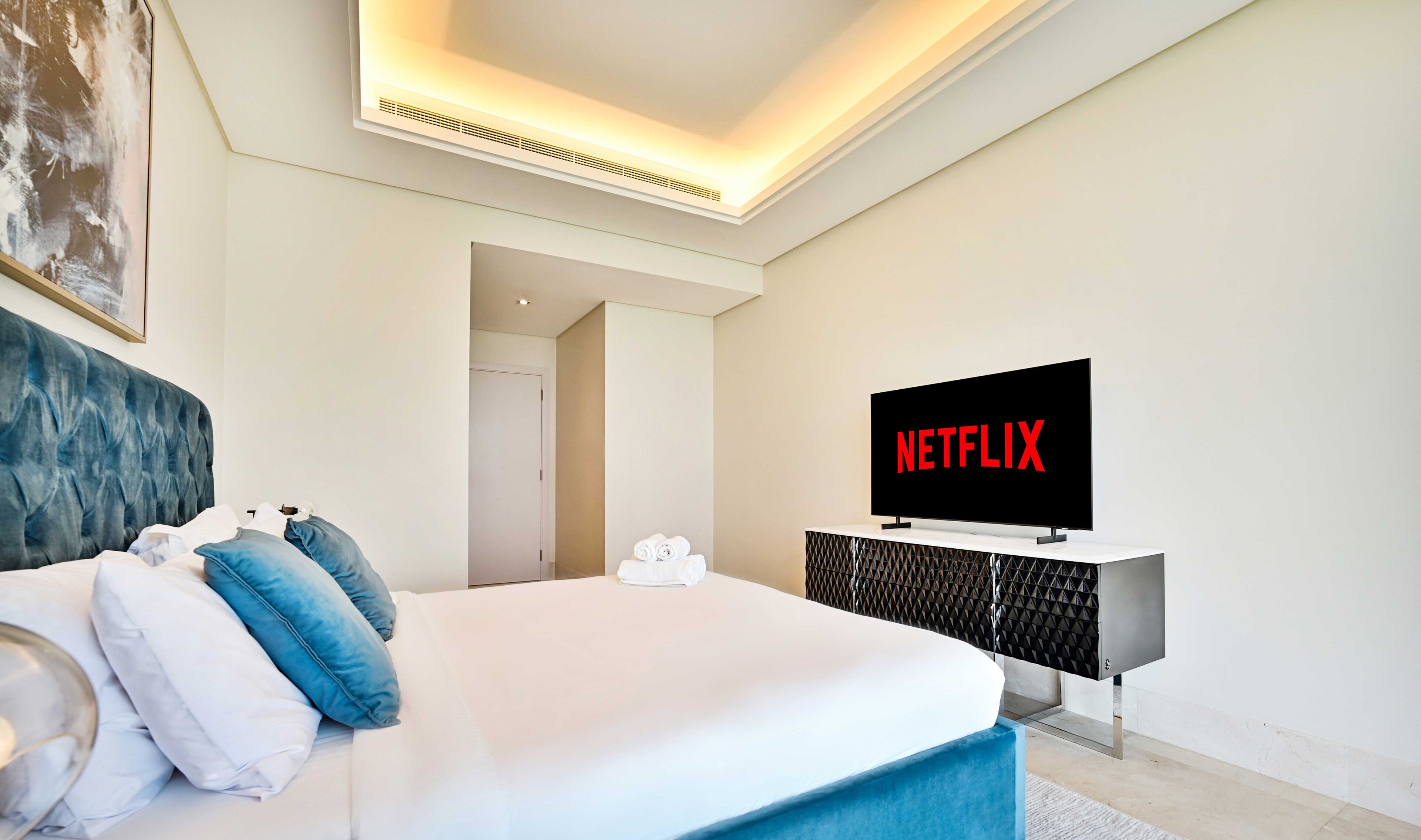 Ein modernes Hotelzimmer mit einem großen Bett, einem Flachbildfernseher, der das Netflix-Logo zeigt, einem weißen Kopfteil und einem minimalistischen Design.