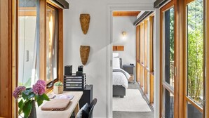Habitación de hotel pequeña con escritorio de madera, cama con ropa de cama blanca y ventanas amplias con vistas a zonas verdes.