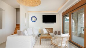 Ein modernes Wohnzimmer mit einem weißen Sofa, einem Holztisch und einem Korbstuhl. Ein Flachbildfernseher ist an der Wand angebracht, und durch eine Glastür ist eine Steinwand zu sehen.