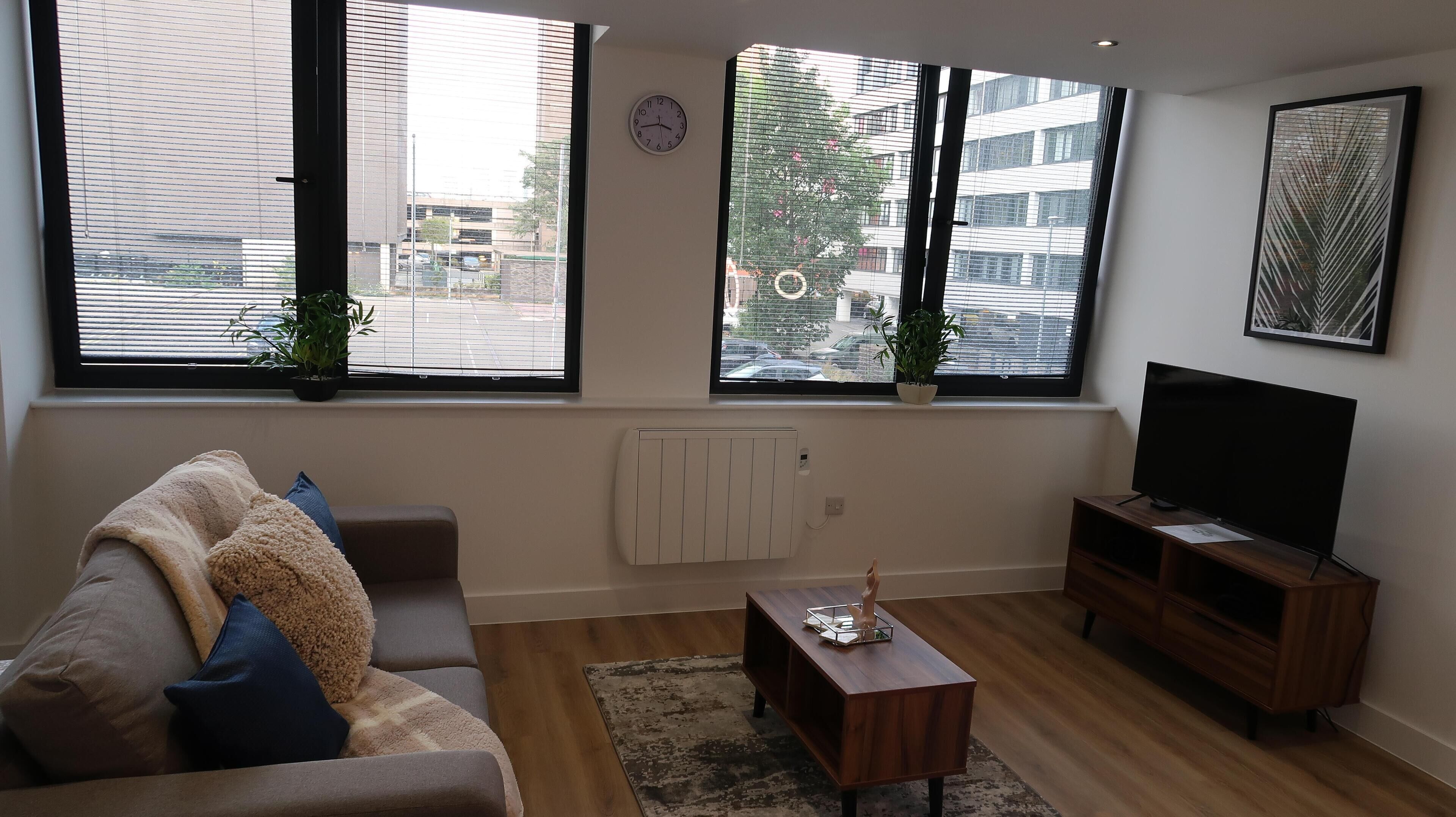 Ein modernes Wohnzimmer mit Sofa, Couchtisch, Fernseher und großen Fenstern mit Jalousien.