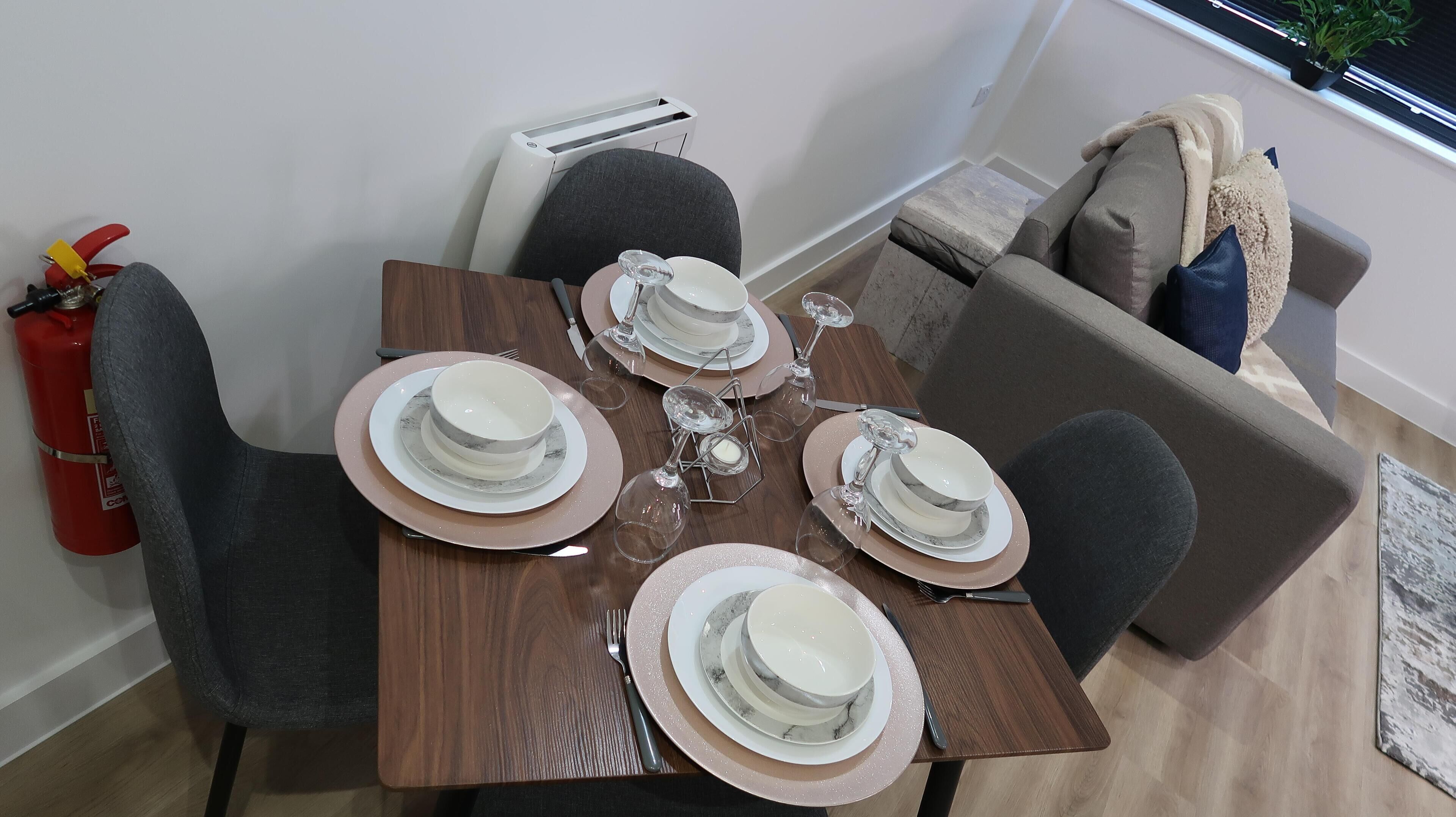 Una mesa de comedor dispuesta con platos, tazones y copas de vino, rodeada de sillas grises y un sofá.