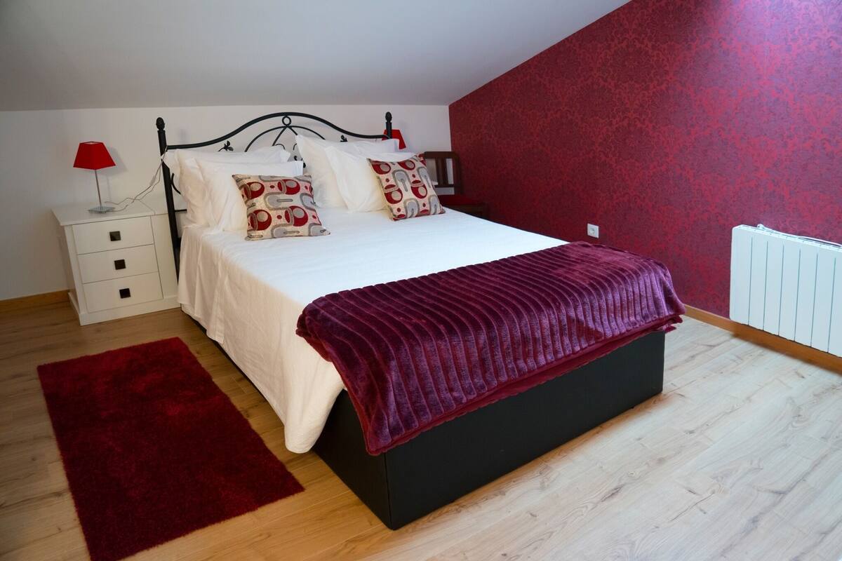 Ein Schlafzimmer mit einem schwarzen Bett, weißer Bettwäsche, einem roten Kissen mit Muster, einem roten Teppich, einem weißen Nachttisch und einer roten Lampe.