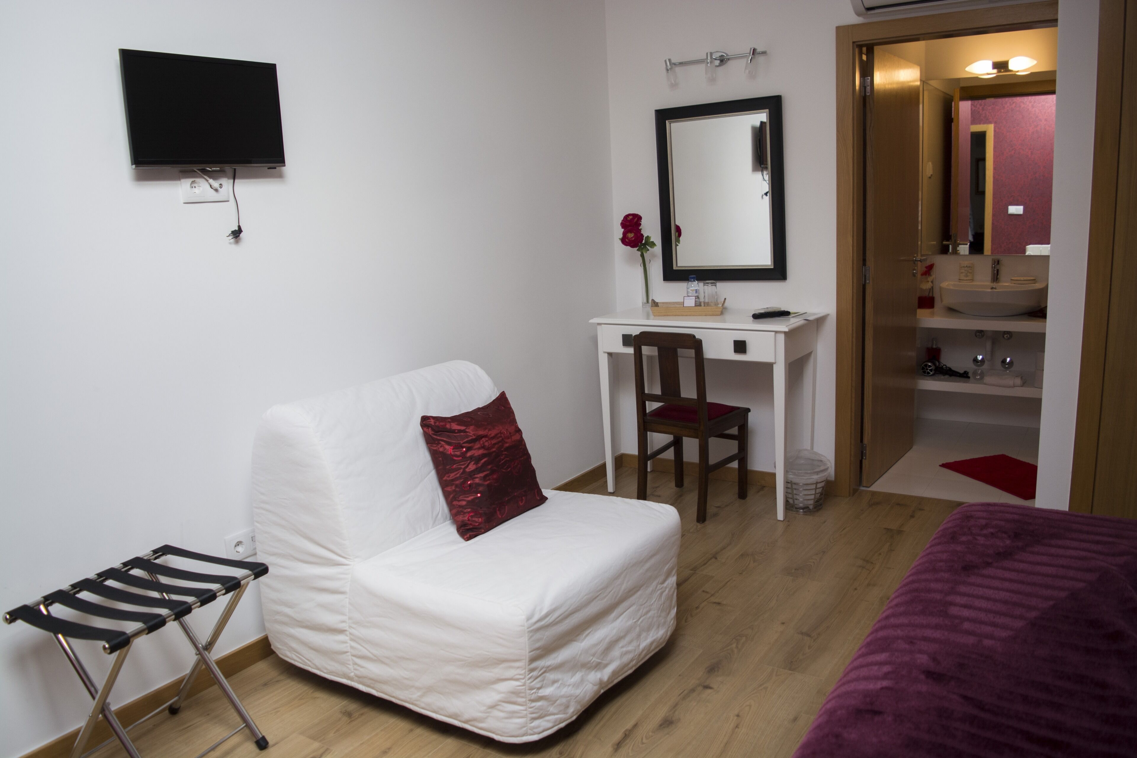 A compact hotel room with a single bed, a small desk with a chair, a television mounted on the wall, and a bathroom visible through an open door.