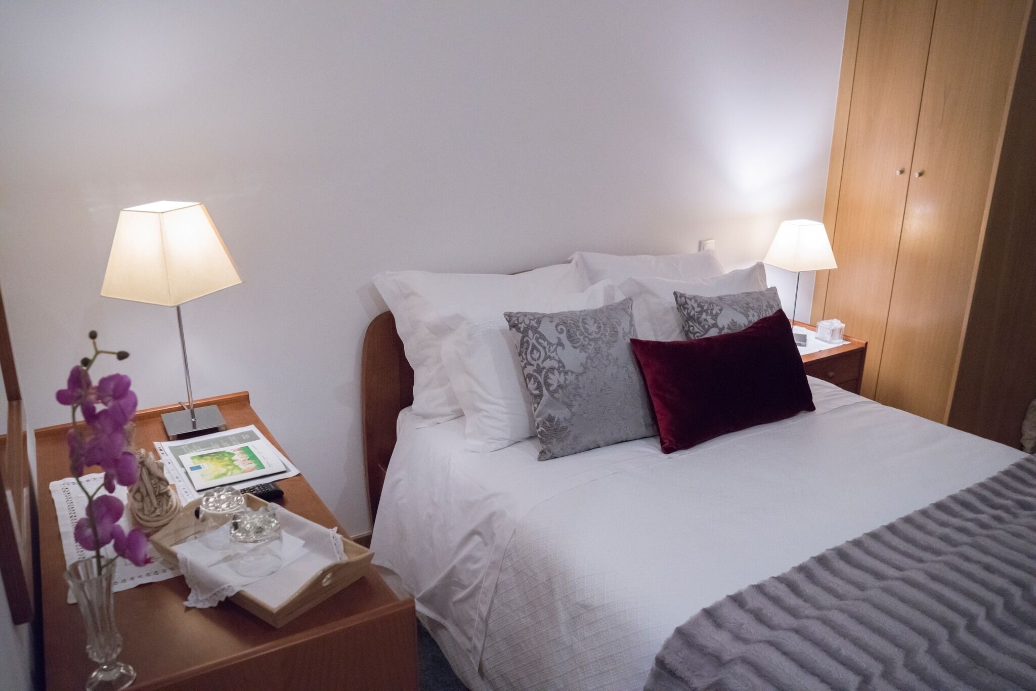 A neatly made bed with white and patterned pillows, a grey blanket, and a bedside table with a lamp, a book, and a vase with flowers.