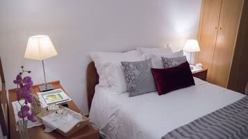 A neatly made bed with white and patterned pillows, a grey blanket, and a bedside table with a lamp, a book, and a vase with flowers.