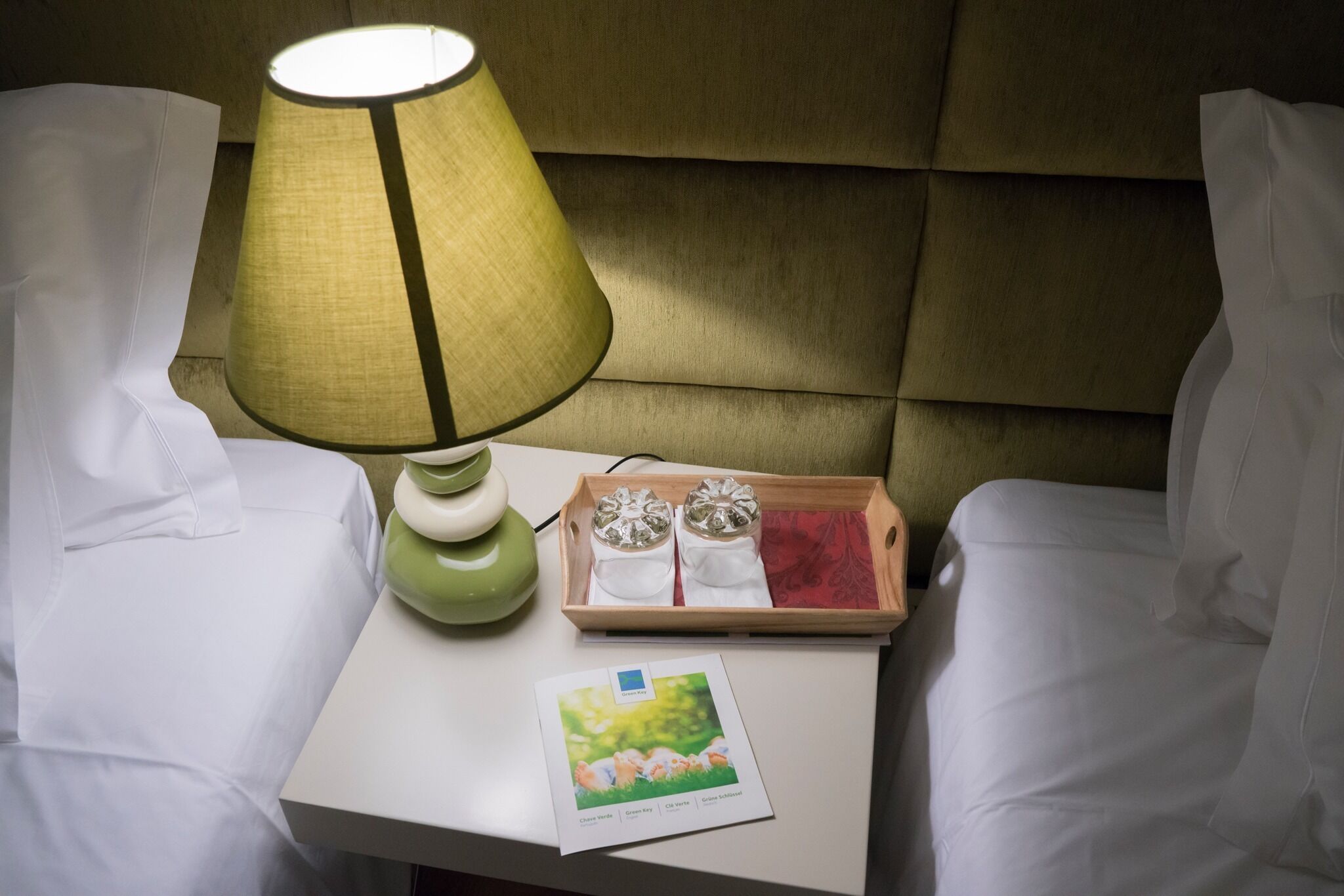 Ein ordentlich bezogenes Bett mit weißen Bettwäsche, eine grüne Lampe, ein Tablett mit zwei weißen Flaschen und eine Karte auf dem Nachttisch.