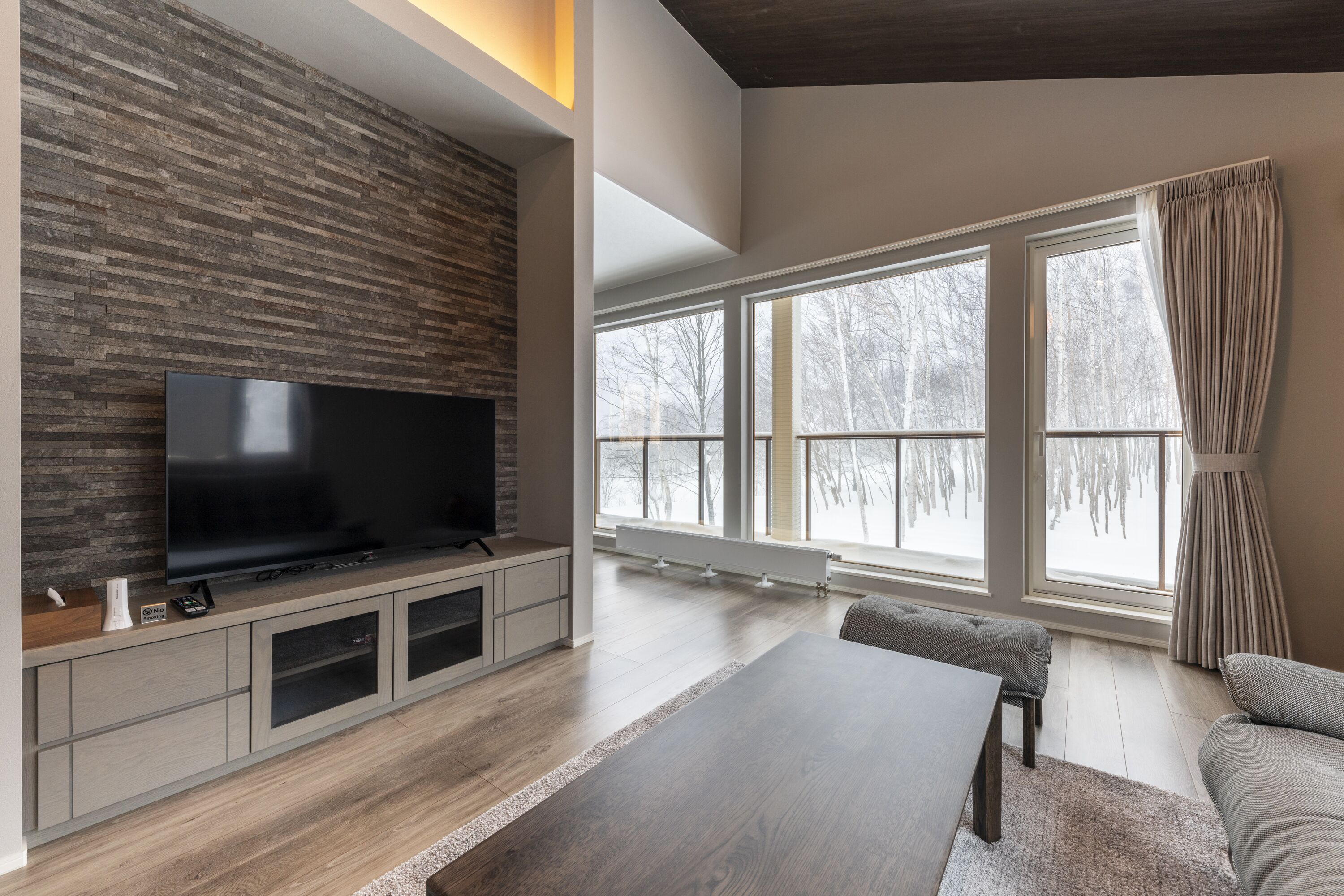 A modern living room with a flat-screen TV, wooden flooring, and large windows with curtains.