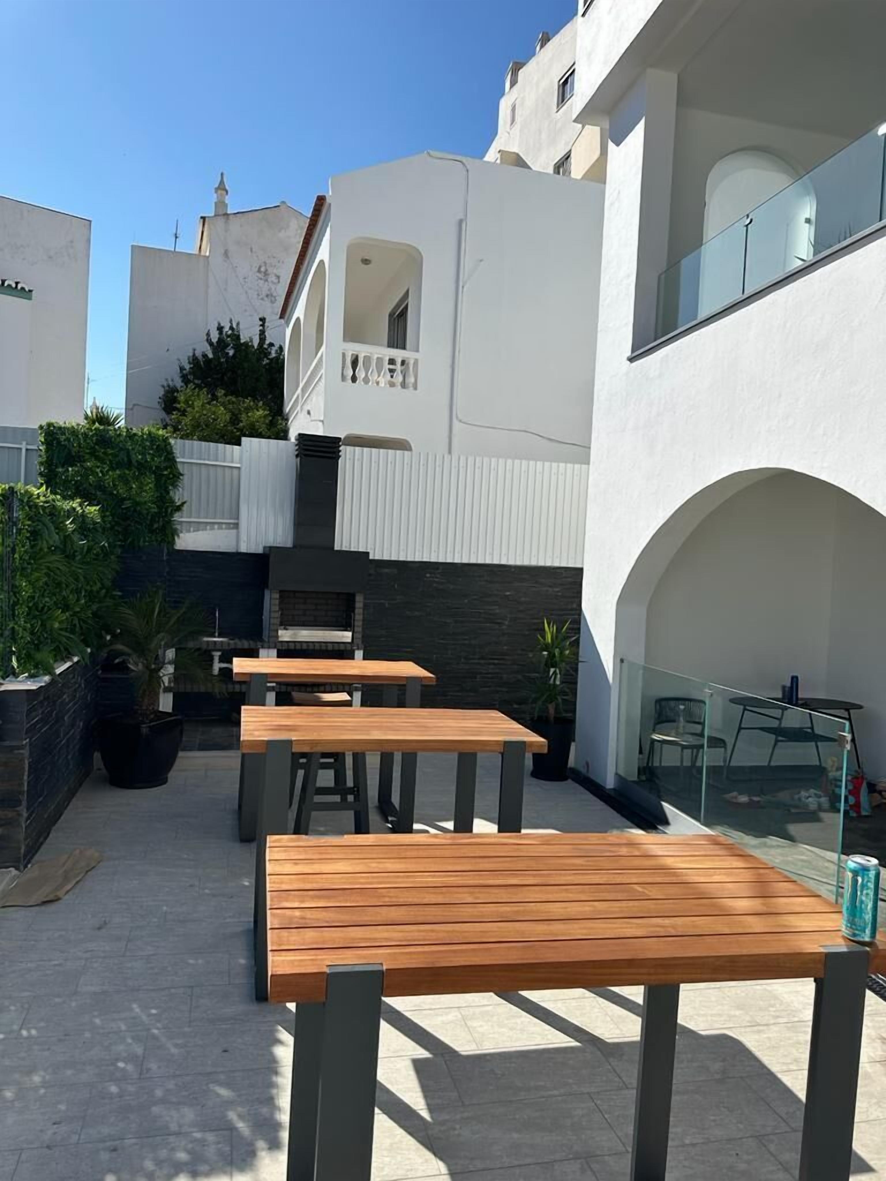 An outdoor patio with wooden tables and chairs, surrounded by white buildings with balconies.