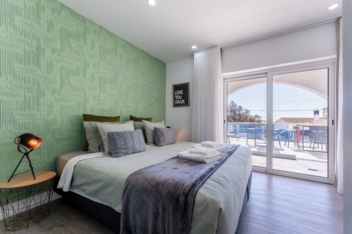 A modern bedroom with a green textured wall, a large bed with multiple pillows, a bedside lamp, and a view of the outdoors through a sliding glass door.