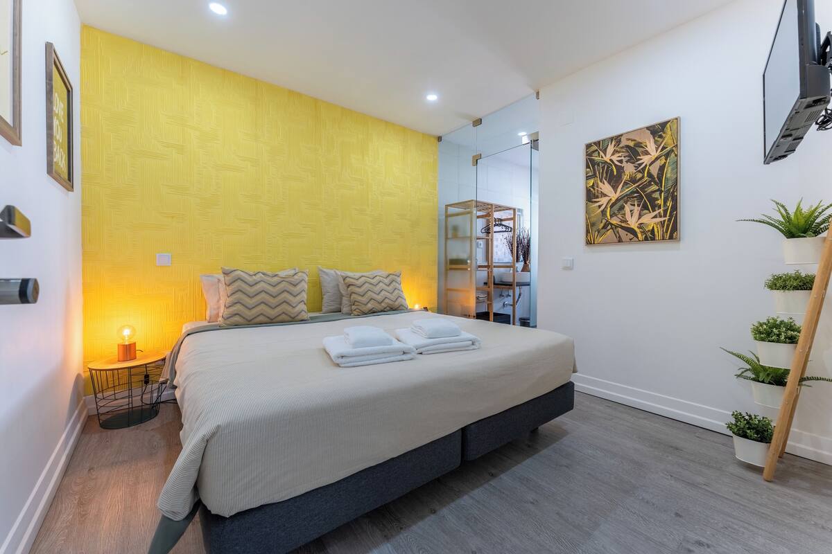A modern bedroom with a large bed, a bedside table with a lamp, a wardrobe, and a wall-mounted TV.