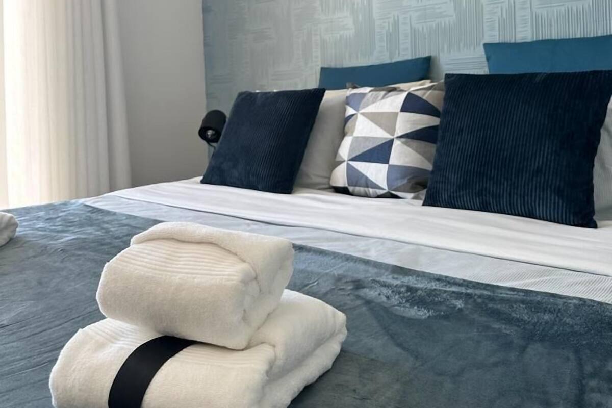A neatly made bed with a blue and white patterned headboard, a pillow with a geometric design, and a stack of folded towels on the bed.