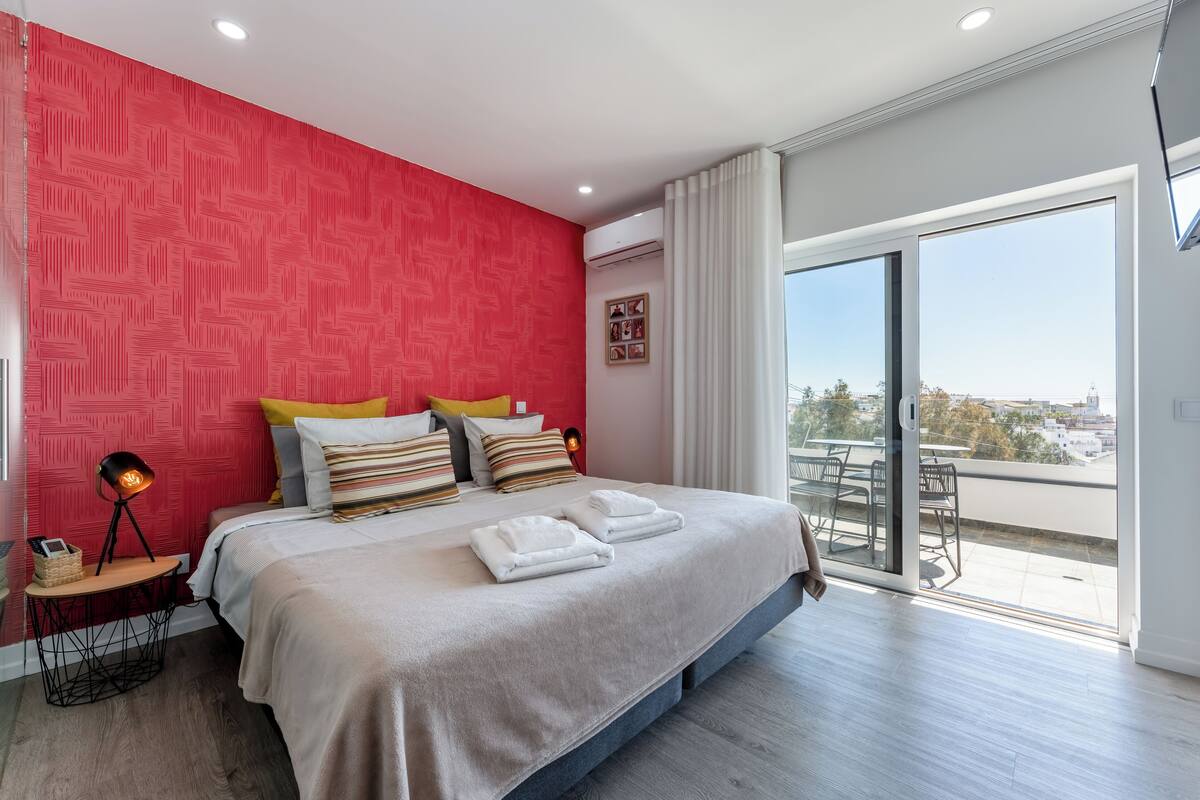 A modern bedroom with a large bed, a red accent wall, and a balcony with a city view.
