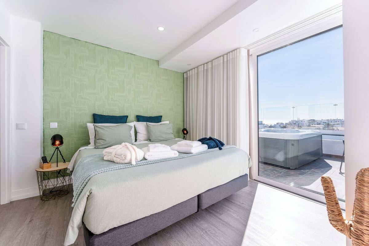 A modern bedroom with a large bed, a nightstand, a lamp, and a view of the city through a large window.