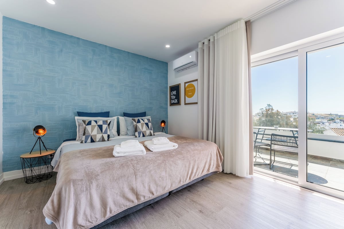 A bedroom with a large bed, a view of the city, and a balcony with a table and chairs.