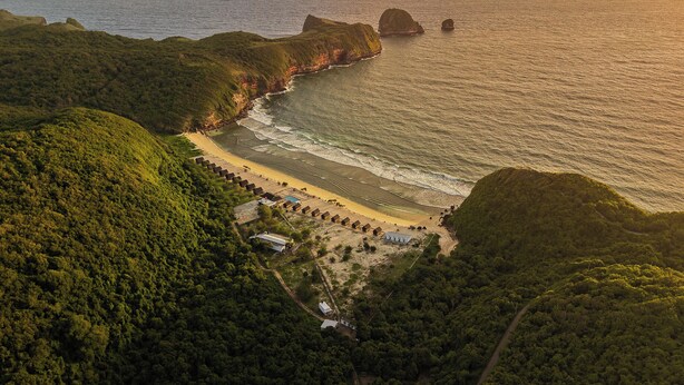 An aerial view of a coastal area with a beach, a row of houses, and a forested hill. - Umarimba Beach Lodge (Mertak)