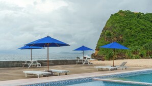 A poolside area with lounge chairs and umbrellas, a view of the ocean, and a rocky cliff. - Umarimba Beach Lodge (Mertak)