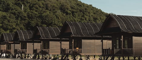 A row of wooden cabins with balconies, set on stilts, against a backdrop of a forested hill.