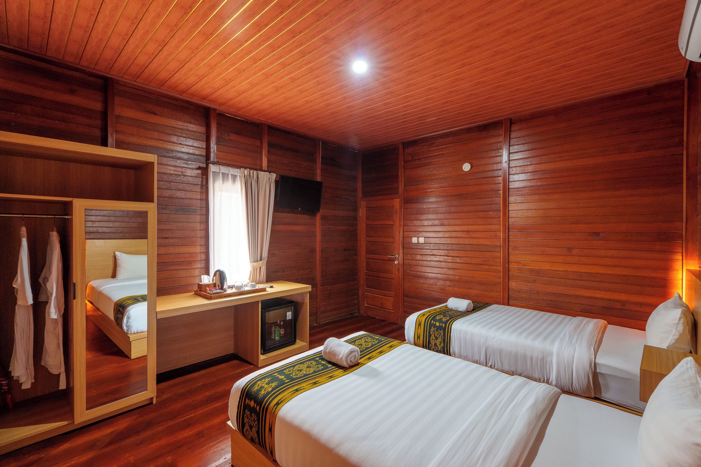 A wooden-paneled room with two beds, a desk, a mirror, and a wardrobe.