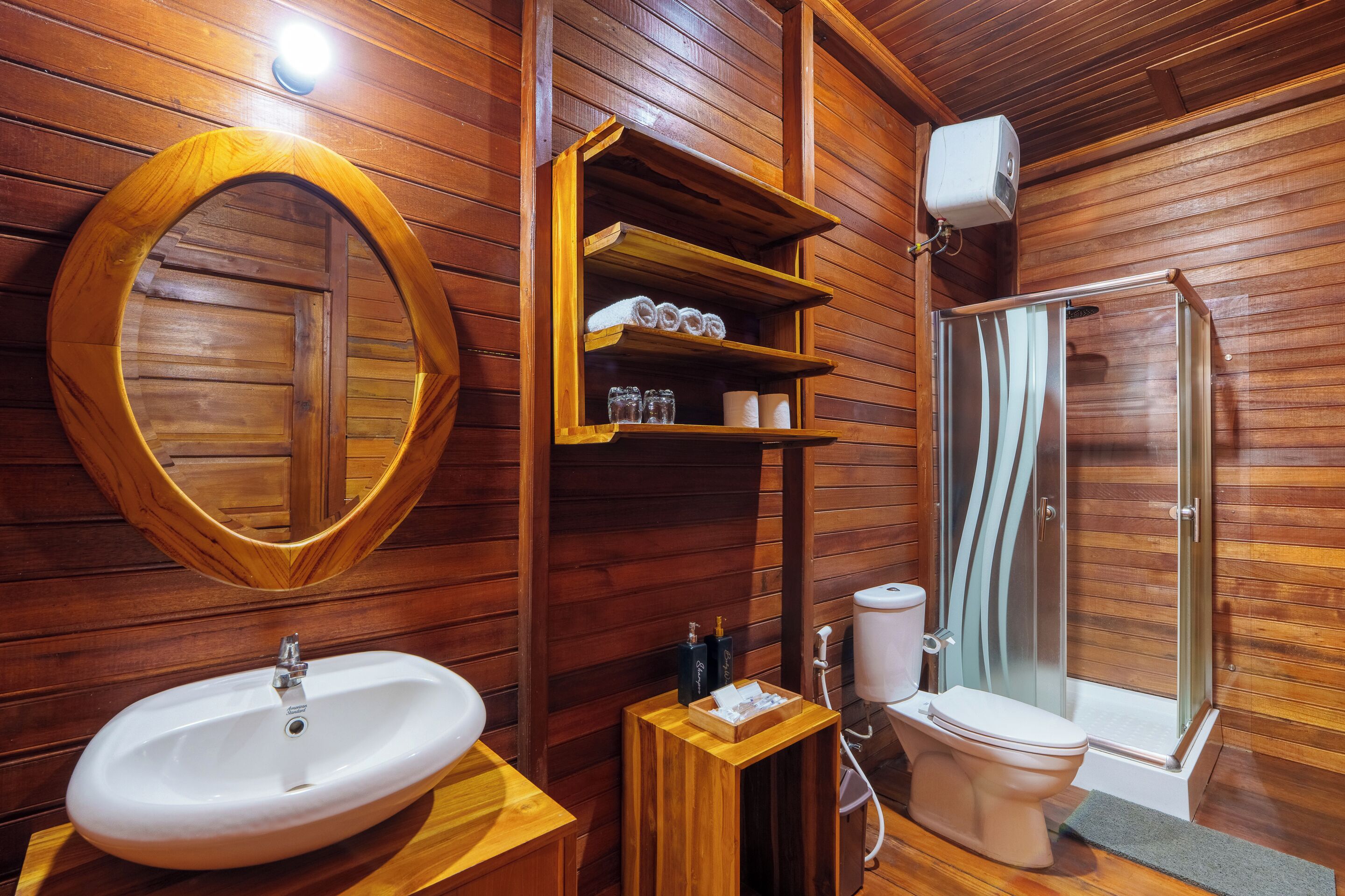 Une salle de bain aux murs lambrissés, équipée d’un lavabo, d’un miroir, d’étagères, de toilettes et d’une douche.
