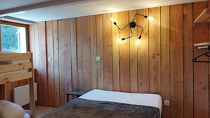 Quarto com paredes de madeira, cama com colchão branco e manta marrom, um banco de madeira e uma pequena mesa.