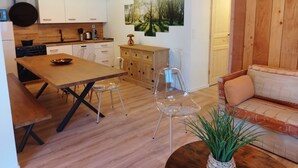 Uma cozinha moderna com armários brancos, uma mesa de jantar de madeira e cadeiras de plástico transparente. Há um banco de madeira e um sofá na sala de estar.