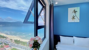 A room with a large window overlooking a beach and ocean, a bed with white bedding, and a vase with flowers. - Ocean View Home Danang (Da Nang)