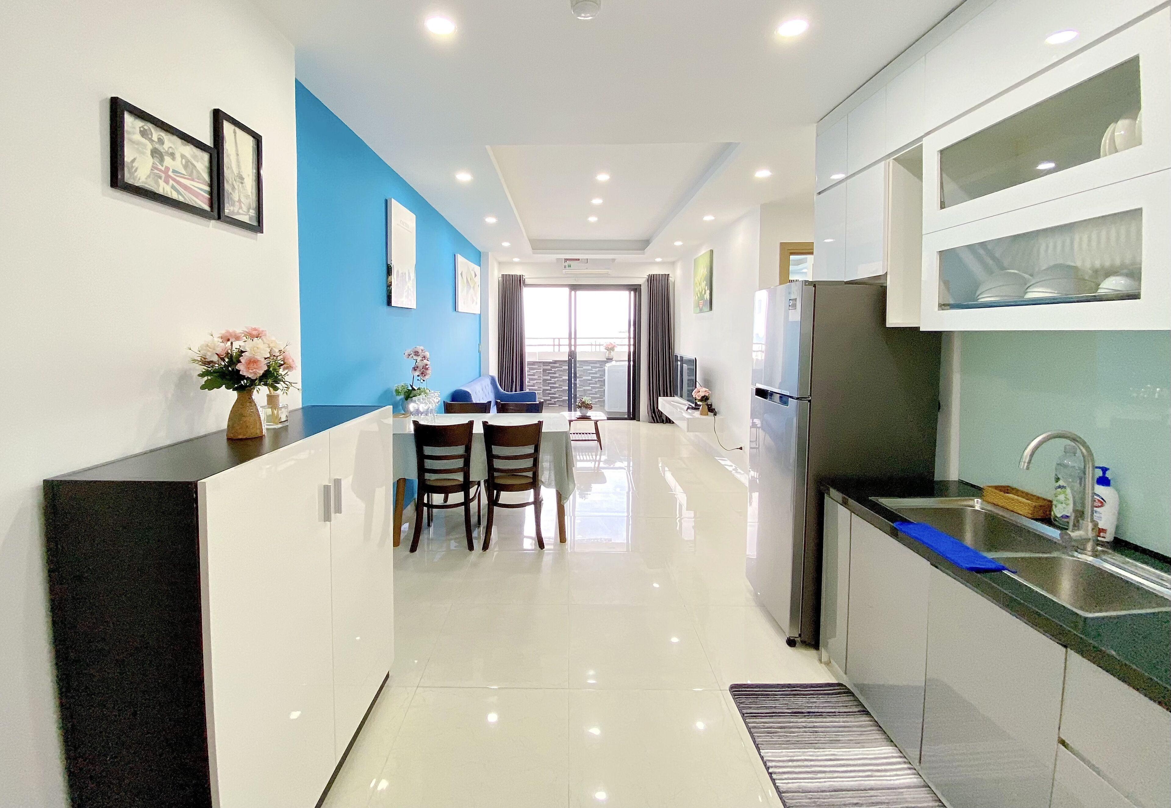 A modern kitchen with white cabinets, a stainless steel refrigerator, and a blue countertop. There is a dining area with two chairs and a table, and a view of a balcony through a glass door.