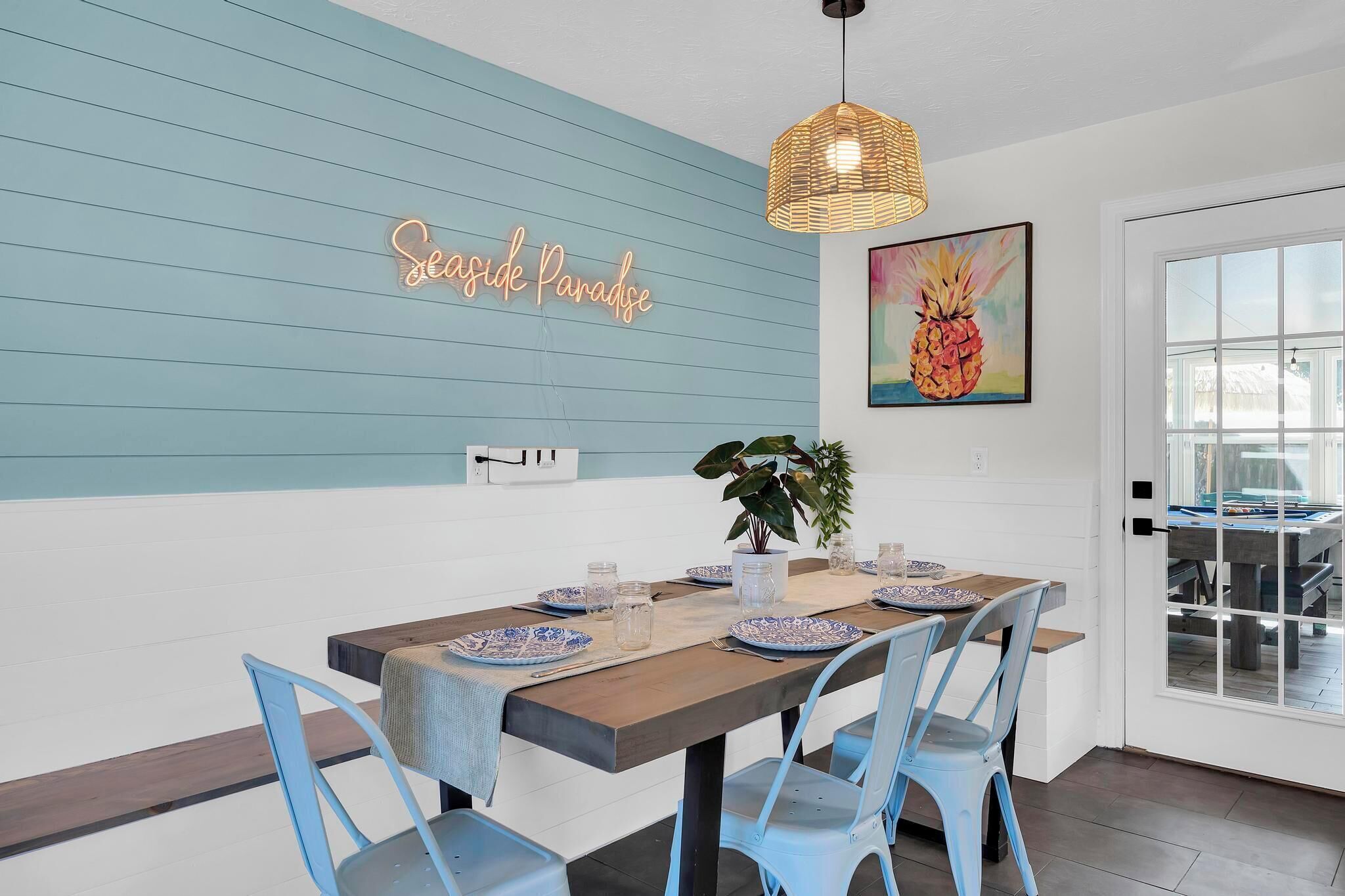 A dining area with a wooden table set for four, blue chairs, a neon sign, and a pineapple painting.