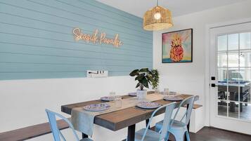 A dining area with a wooden table set for four, blue chairs, a neon sign, and a pineapple painting.