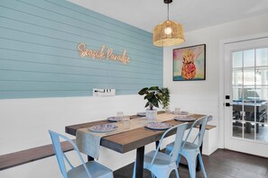A dining area with a wooden table set for four, blue chairs, a neon sign, and a pineapple painting. - Seaside Paradise: Dive into Luxury with Saltwater Pool, Spa, and Chemical-Free Bliss (Ponte Vedra Beach)