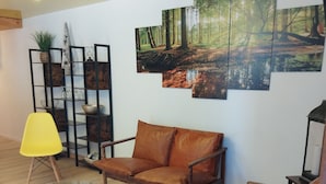 A modern living room with a brown leather sofa, a glass coffee table, and a yellow chair. There is a wooden floor and a wall with a forest-themed painting.