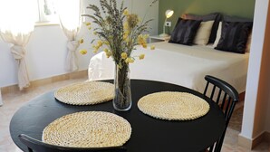 Une table ronde surmontée de sets de table en fibres tressées, un vase transparent contenant des fleurs séchées et, au fond, un lit agrémenté de coussins.