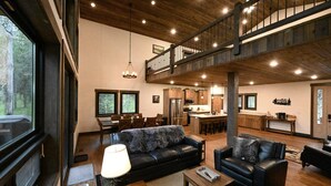 A spacious living area with a large black leather sofa, a wooden coffee table, and a dining table with chairs. A kitchen with modern appliances is visible in the background.