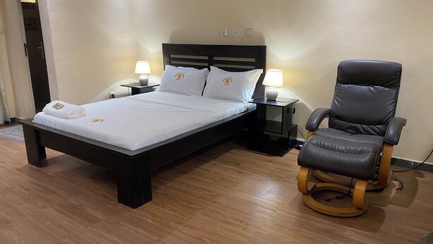 A neatly made bed with white linens and two pillows, a wooden headboard, a black nightstand with a lamp, and a leather chair with a wooden base.