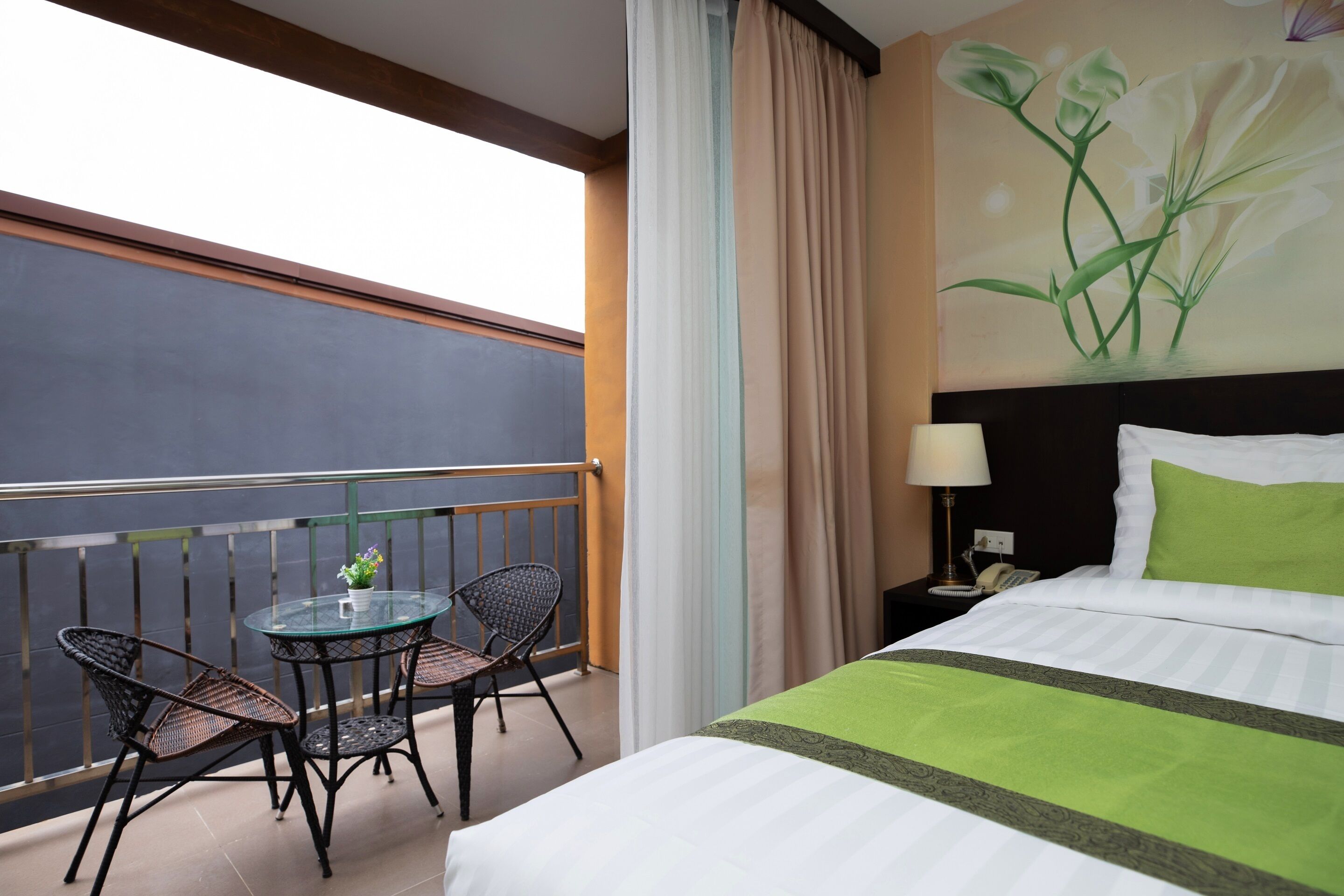 Habitación de hotel con balcón, cama con ropa de cama verde, mesita de noche con lámpara y un cuadro de flores en la pared.