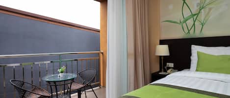 Habitación de hotel con balcón, cama con ropa de cama verde, mesita de noche con lámpara y un cuadro de flores en la pared.
