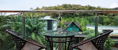 Un balcón con sillas de mimbre y una mesa de vidrio, con vistas a un paisaje verde exuberante y edificios.