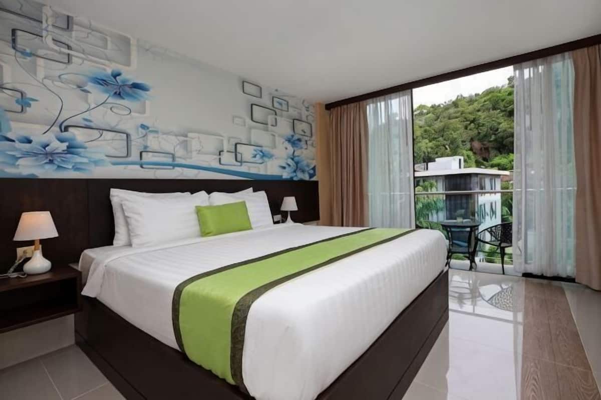 A hotel room with a large bed, a nightstand, a wall with abstract art, a balcony with a view of greenery, and a table with chairs.