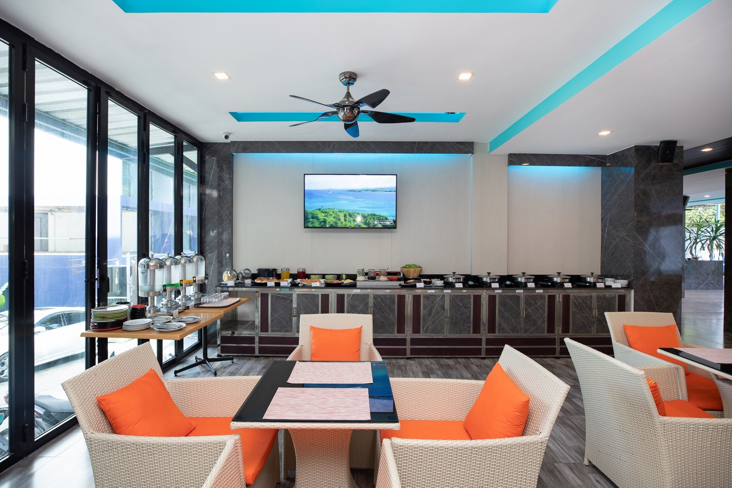 Un espace repas moderne, doté de meubles en osier, d’un ventilateur de plafond et d’un écran de télévision diffusant un paysage pittoresque.