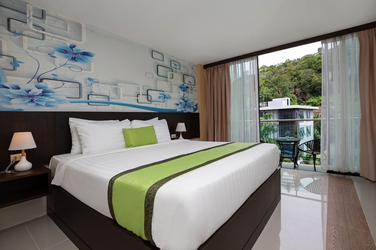 A hotel room with a large bed, a nightstand with a lamp, a wall with a floral mural, and a balcony with a table and chairs.