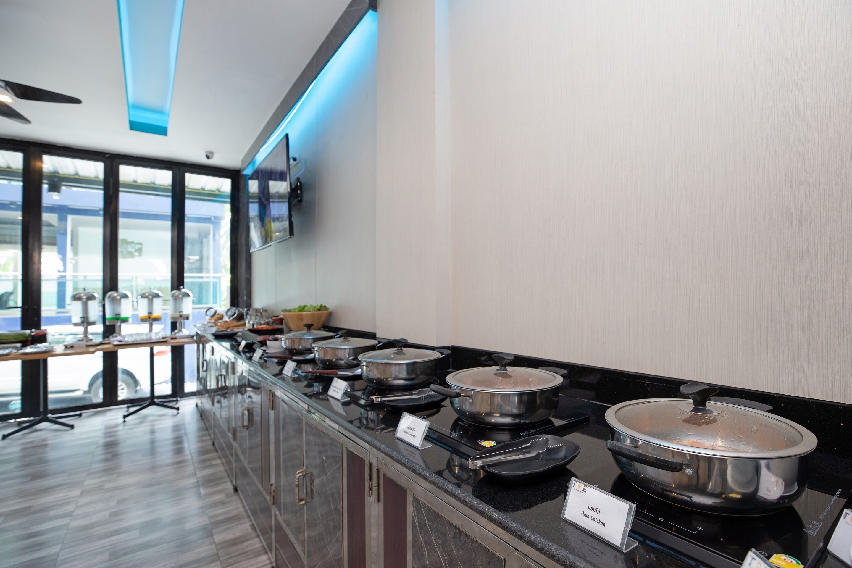 A buffet setup with multiple hot food stations, a ceiling fan, and a view of the outdoors through large windows.