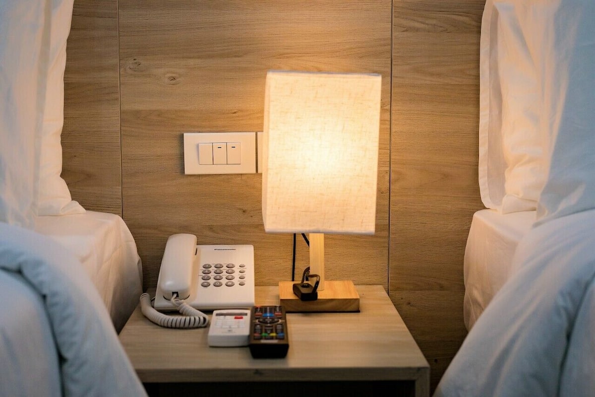 A hotel room with a wooden wall, a bedside lamp, a telephone, and a remote control on the nightstand.