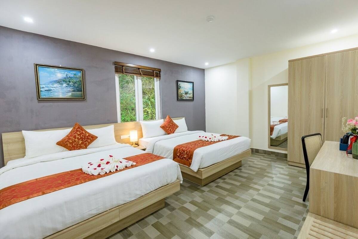 A hotel room with two beds, a wooden wardrobe, a desk with a chair, and a window with a view of greenery.