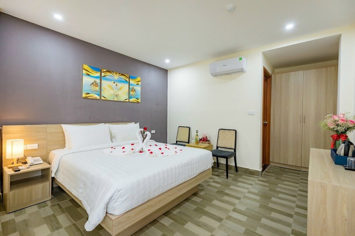 A hotel room with a large bed, a nightstand with a lamp, two chairs, a small table with a vase of flowers, and artwork on the wall.