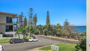 Rua residencial com dois carros estacionados, céu azul límpido e vista para o mar.
