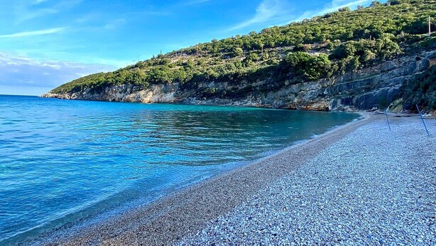 A pebble beach with clear blue water and a rocky hillside with sparse vegetation.